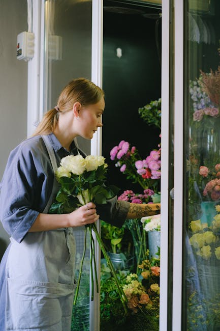 A young woman, with light brown hair tied back, is selecting a bouquet of fresh white roses at a florist shop for flower delivery. She is wearing a grey apron over dark clothing and is holding the bou