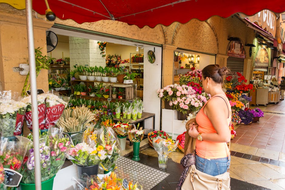 A woman with her hair tied back, wearing an orange sleeveless top and blue jeans, stands outside a flower shop on Castellain Road Maida Vale. She is holding a brown coat and a beige bag while examinin