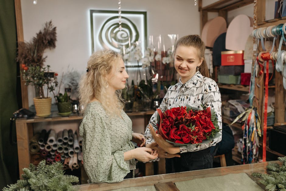 A young woman with light brown hair tied in a high ponytail, smiling as she presents a vibrant bouquet of red roses, lilies, and greenery wrapped in brown paper, to a blonde woman with curly hair wear