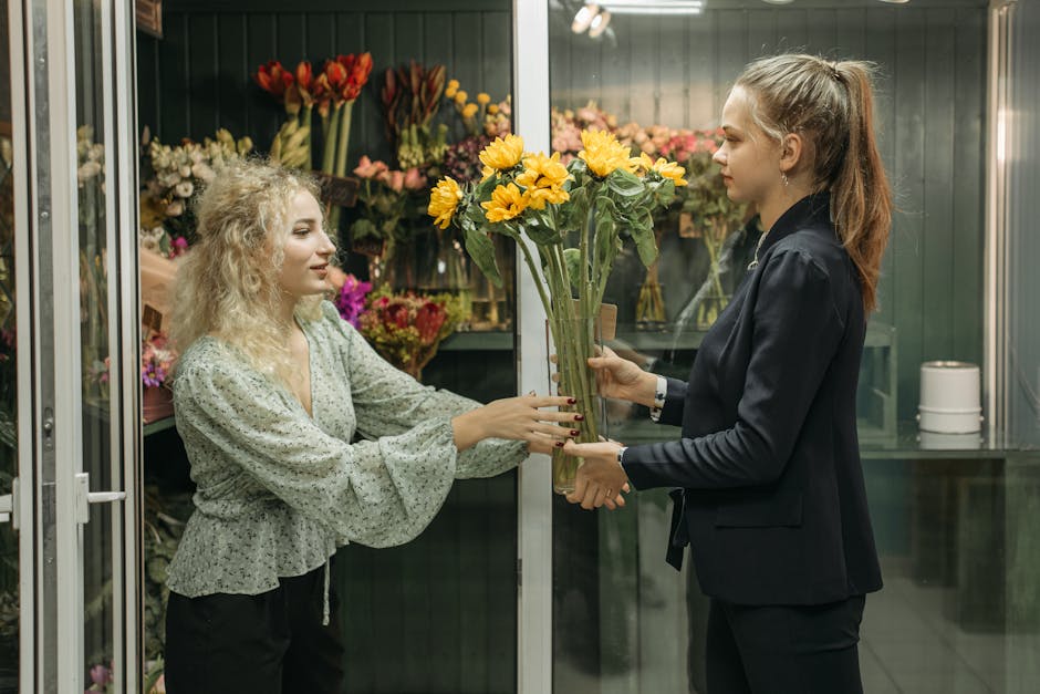 A florist with light blonde, curly hair wearing a light floral blouse is handing a bouquet of bright yellow sunflowers wrapped in clear cellophane to a customer in a flower shop. The customer, with lo