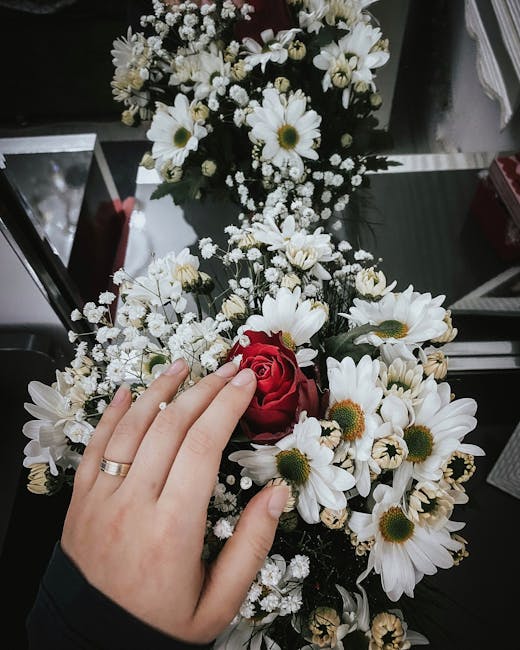 A hand with a silver wedding band gently resting on a floral bouquet consisting of white daisies with yellow centers, small white baby's breath, and a single red rose at the center. The bouquet is arr