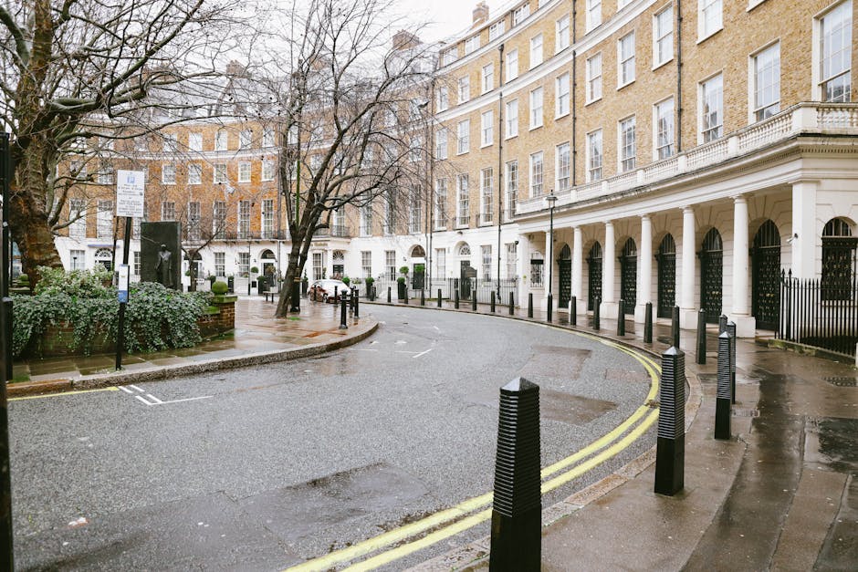 The image depicts a quiet street corner in Maida Vale, featuring a historic row of white stucco-fronted Georgian buildings with large sash windows and decorative columns along the pavement. The street