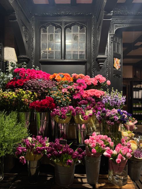 A display of vibrant, fresh flowers arranged in metal buckets and vases within an elegant, dark-toned interior featuring intricate wooden paneling and a large, decorative window. The bouquet mixture i