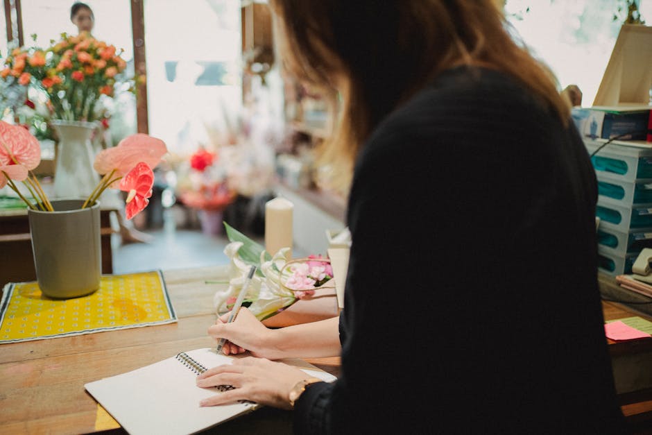 A florist working at a wooden table with a bouquet of pink roses, greenery, and decorative wrapping materials, preparing a floral gift in a bright, well-lit flower shop. The bouquet is partially wrapp