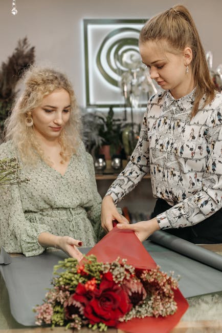 A florist from flowersmaidavale.co.uk is preparing a floral gift, arranging a bouquet featuring vibrant red roses, pink baby's breath, and green foliage, all carefully wrapped in red floral paper. The