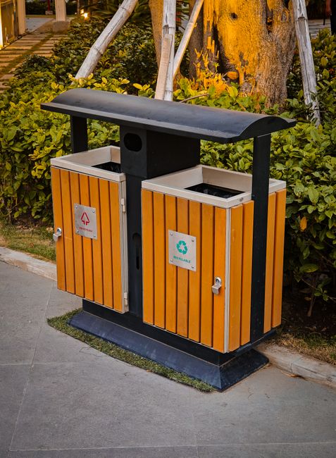 A double-chamber outdoor waste disposal bin with two separate compartments, each with orange wooden slatted doors and metal handles. The left side has a red recycling symbol indicating general waste, 