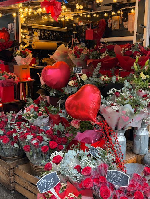 An outdoor floral display featuring a variety of fresh flowers, predominantly red and white roses, arranged in baskets and wrapped bouquets. The arrangement includes silver-green foliage and heart-sha