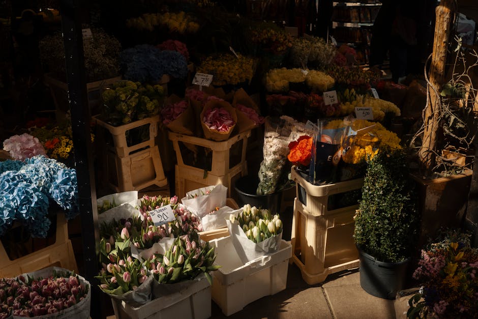 A vibrant flower stall showcasing a diverse selection of fresh flowers arranged in baskets and crates. In the foreground, pink and white tulips are wrapped in white paper, with some priced at £2.50, 