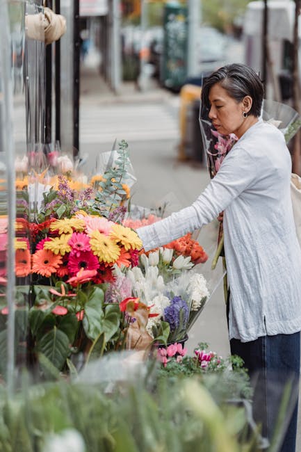 A woman with short dark hair, wearing a light grey cardigan over a white shirt, is selecting a vibrant bouquet of fresh flowers from a flower stall on a city sidewalk. The bouquet features a mix of br