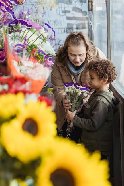 A woman and a young girl are inside a bright flower shop, browsing and selecting floral gifts. The woman, with curly blonde hair and a light brown jacket, carefully presents a bouquet of mixed purple 